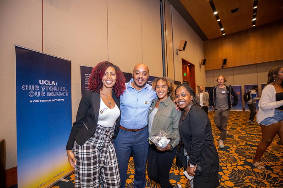Three female and one male guests standing in front of UCLA Centennial banner and booth at a Black Bruin resourceevent